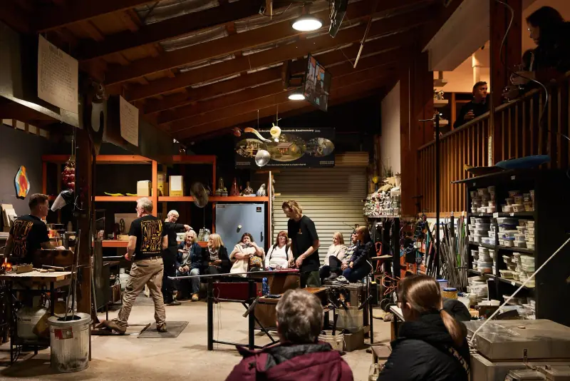Visitors seated around the hot shop floor watching the glassblowers work on a Friday night