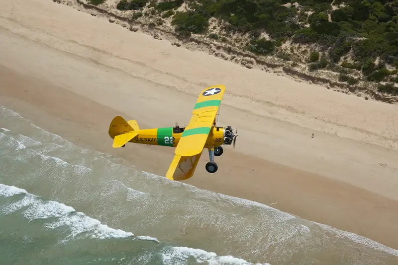 A yellow open-cockpit biplane flying low over the surf and a sandy beach