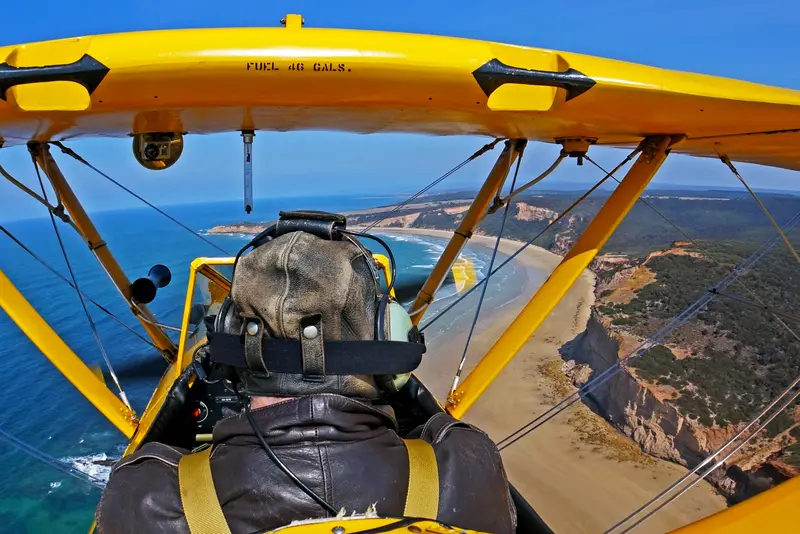 The view from the open cockpit, past the pilot's head and the yellow wings to the coastline below